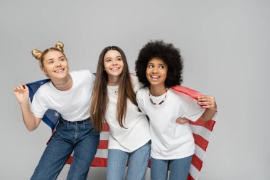 Beyaz tişörtlü ve kot pantolonlu genç ve çok ırklı kız arkadaşlar Amerikan bayrağını tutarken gülümsüyor ve gri arka planda duruyorlar enerjik genç arkadaşlar vakit geçiriyorlar, arkadaşlıklar