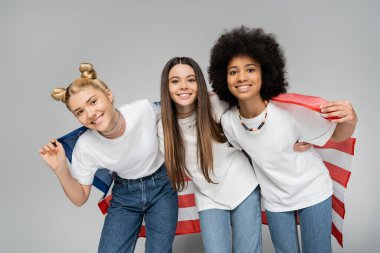Beyaz tişörtlü, kot pantolonlu, gri arka planda Amerikan bayrağıyla poz veren pozitif etnik kökenli genç kızlar, enerjik genç arkadaşlar zaman geçiriyorlar, arkadaşlıklar...