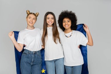 Beyaz tişörtlü ve kot pantolonlu neşeli ve etnik çeşitlilikte genç kızlar Avrupa bayrağını bir arada tutuyorlar ve gri, enerjik genç arkadaşların üzerinde izole bir şekilde duruyorlar. 
