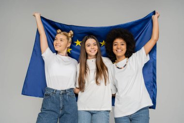 Beyaz tişörtlü ve kot pantolonlu pozitif ve etnik çeşitlilikte genç kızlar, gri, enerjik genç arkadaşlarıyla vakit geçirirken ellerinde mavi Avrupa bayrağı tutuyorlar.
