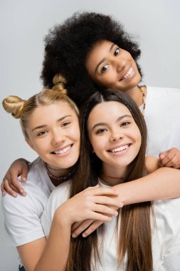 Portrait of smiling multiethnic teen friends hugging brunette girlfriend in white t-shirt and looking at camera together isolated on grey, energetic teenage friends spending time clipart
