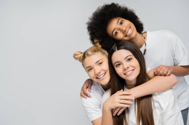 Gleeful and teenage girls in white t-shirts hugging each other and looking at camera while standing isolated on grey, energetic teenage friends spending time, copy space, friendship and companionship clipart