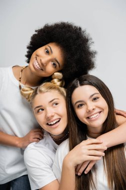 Portrait of smiling african american teenage girl in white t-shirt hugging blonde and brunette girlfriends while standing isolated on grey, energetic teenage friends spending time clipart