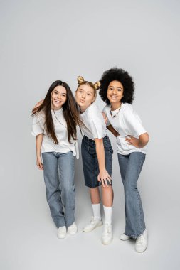 Full length of stylish and multiethnic teenage girlfriends in white t-shirts and jeans posing and looking at camera on grey background, adolescence models and generation z concept clipart