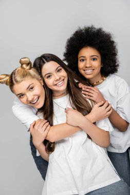 Kotlu ve beyaz tişörtlü neşeli ve çok ırklı genç kız arkadaşların portresi esmer arkadaşa sarılıp gri, ergenlik modelleri ve Z jenerasyonunda izole edilmiş poz veriyorlar.