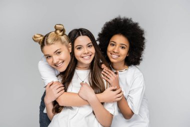 Portrait of positive and interracial teenage girls in white t-shirts hugging brunette girlfriend and looking at camera isolated on grey, adolescence models and generation z concept clipart