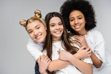 Portrait of cheerful and multiethnic teenage girls in white t-shirts hugging each other and looking at camera while standing isolated on grey, teenage friends having fun together clipart