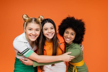 Portrait of positive and interracial teenage girlfriends in trendy outfits hugging each other and looking at camera while standing isolated on orange, trendy generation z concept clipart