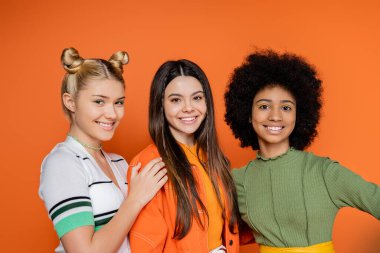 Portrait of positive and multiethnic teenage girlfriends in stylish outfits with makeup looking at camera while standing isolated on orange, trendy generation z concept, friendship and companionship clipart