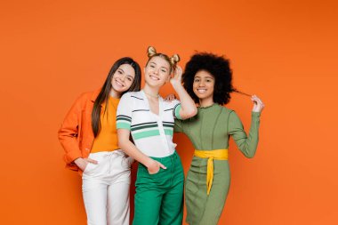 Makyajlı çok ırklı ve genç kız arkadaşlar turuncu arka plandaki kameraya, kültürel çeşitliliğe ve Z kuşağı moda konseptine, arkadaşlığa bakıyorlar. 