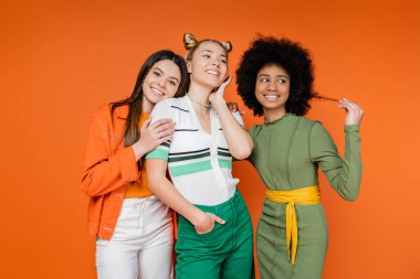 Cesur makyajlı gülümseyen çok ırklı genç kızlar moda sarışın kız arkadaşının yanında poz veriyorlar ve turuncu arka plan, kültürel çeşitlilik ve Z kuşağı moda konsepti üzerinde birlikte duruyorlar.