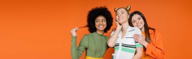 Cheerful interracial teen girls in trendy outfits and makeup posing near blonde girlfriend while standing isolated on orange, cultural diversity and generation z fashion concept, banner  clipart