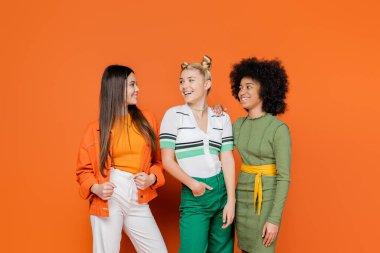 Moda kıyafetleri içinde neşeli ve çok ırklı kız arkadaşlar turuncu arka plan, kültürel çeşitlilik ve Z kuşağı moda konsepti üzerinde yan yana durup konuşuyorlar ve birbirlerine bakıyorlar.