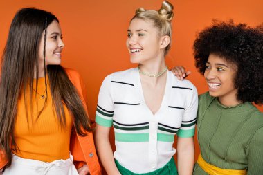 Pozitif ve çok ırklı genç kız arkadaş portresi moda kıyafetleri içinde makyajlı turuncu arka plan, kültürel çeşitlilik ve Z kuşağı moda konsepti hakkında konuşuyor ve birbirlerine bakıyorlar.