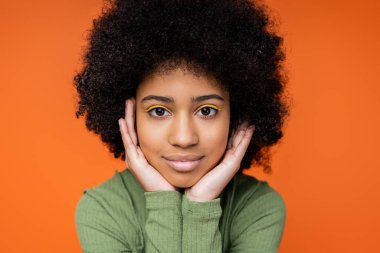 Portrait of teenage african american girl with bright makeup wearing green dress and looking at camera while touching cheeks isolated on orange, youth culture and generation z concept  clipart