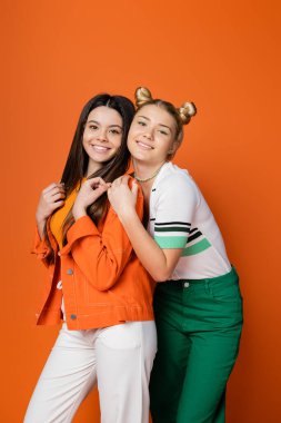 Cesur makyajlı neşeli sarışın kız günlük kıyafetli modaya uygun esmer kız arkadaşına sarılıyor ve turuncu renkli, modaya uygun, tarz, arkadaşlık ve bağlılık duygusu olan kızlara bakıyor.