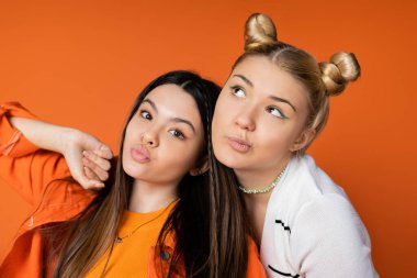 Portrait of stylish blonde teenage girl with bold makeup posing and standing near brunette girlfriend pouting lips isolated on orange, fashionable girls with sense of style clipart