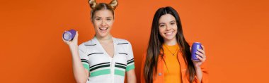 Joyful and stylish blonde and brunette teenage models looking at camera while holding drink in tin cans on orange background, fashionable girls with sense of style, banner  clipart