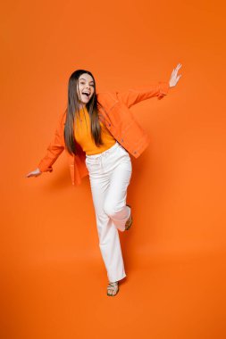 Full length of excited and trendy brunette teen model in denim jacket and pants posing and looking at camera while standing on orange background, cool and confident teenage girl clipart