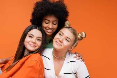 Smiling african american teen model with bright makeup hugging fashionable girlfriends and posing together isolated on orange, cool and confident teenage girls, diverse races  clipart