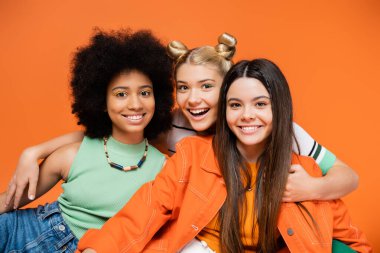 Neşeli sarışın genç kız şık ve çok ırklı kız arkadaşlara cesur makyajlarıyla sarılıp turuncu, havalı ve kendine güvenen çok kültürlü genç kızlar, farklı ırklar 