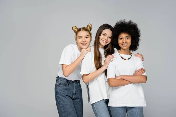 stock image Positive and teenage girls in white t-shirts and jeans hugging confident african american girlfriend crossing arms while standing isolated on grey, lively teenage girls concept, friendship and bonding