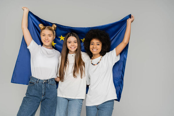 Joyful and multiethnic teenage girls in t-shirts and jeans holding european flag while posing and standing isolated on grey, energetic teenage friends spending time, friendship and companionship