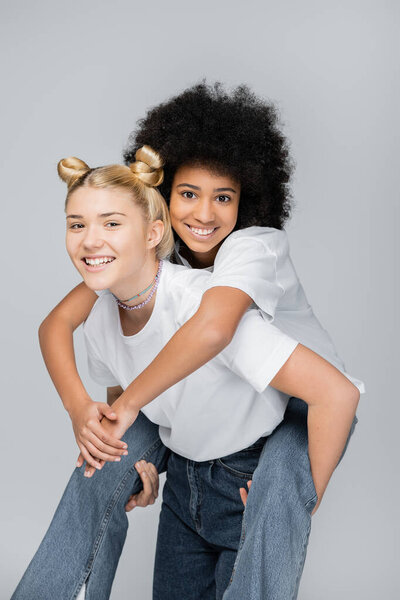 Teen african american girl in jeans and white t-shirt hugging blonde girlfriend while looking at camera and standing isolated on grey, energetic teenage models spending time