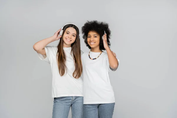 stock image Joyful and interracial teen girls in white t-shirts and jeans listening music in headphones and looking at camera isolated on grey, energetic teenage models spending time, friendship and companionship
