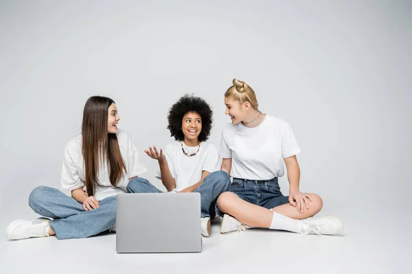 陽気なアフリカ系アメリカ人の女の子でホワイトTシャツとジーンズと話を十代の女の子に一緒に座っている間にノートパソコンの近くにグレーの背景に ティーンボンディング上の共通の関心 — ストック写真