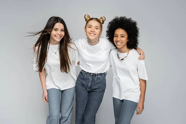 stock image Smiling blonde teen girl in white t-shirt and jeans hugging multiethnic girlfriends and looking at camera on grey background, adolescence models and generation z concept