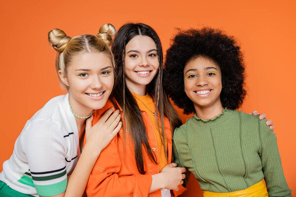 Портрет модных и мультиэтнических девочек-подростков в модных нарядах и макияже, обнимающих и смотрящих в камеру, позируя на оранжевом фоне, модное поколение z концепция
