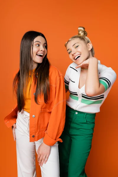 stock image Cheerful and trendy brunette teenager with bright makeup looking at blonde friend holding hand near face while posing on orange background, fashionable girls with sense of style