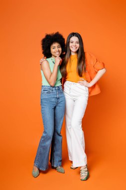 Şık kıyafetli neşeli esmer kız turuncu arka planda parlak makyajlı Afro-Amerikan kız arkadaşına sarılıyor. Kusursuz stil konsepti olan ergen modacılar.