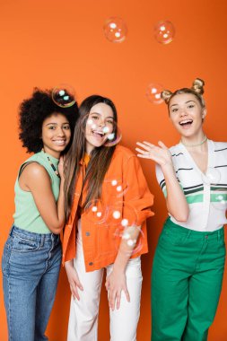 Soap bubbles near positive and interracial teenage girlfriends in casual outfits looking at camera on orange background, teen fashionistas with impeccable style concept clipart