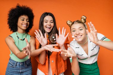 Cheerful and multiethnic teen girlfriends in stylish casual clothes looking at soap bubbles while standing on orange background, teen fashionistas with impeccable style concept clipart