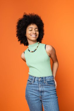 Joyful and stylish african american teenage girl with necklace and bold makeup closing eyes while standing isolated on orange, trendy teenage girl expressing individuality clipart