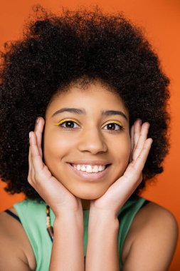 Portrait of positive and stylish african american teenager with bold makeup touching face and looking at camera while posing isolated on orange, trendy teenage girl expressing individuality  clipart