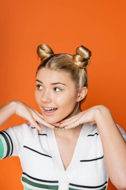 Cheerful and blonde teenager with colorful makeup and hairstyle holding hands near chin and looking away while standing isolated on orange, trendy teenage girl expressing individuality clipart