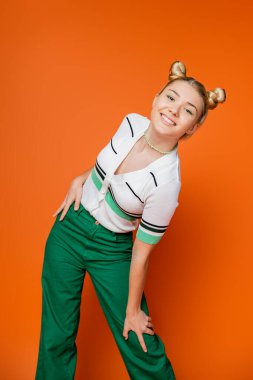 Positive and fashionable teenage girl with hairstyle and bold makeup posing in casual outfit and looking at camera while standing on orange background, fashionable and trendy clothes clipart