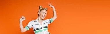 Excited and cheerful blonde teenager with hairstyle and bold makeup posing in casual clothes and looking at camera while standing isolated on orange, fashionable and trendy clothes, banner  clipart