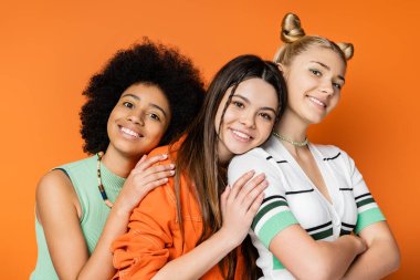 Renkli makyajlı pozitif ve etnik çeşitlilikte genç kızlar sarılıp kucaklaşan sarışın arkadaşlar turuncu, şık ve modaya uygun kıyafetlerle izole edilmiş kameralara bakıyorlar.