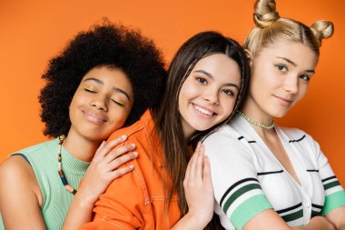 Renkli makyajlı, turuncu, şık ve modaya uygun giysiler içinde tek başına dikilirken kendine güvenen sarışın arkadaşına sarılan pozitif ırklar arası genç kız arkadaşlarının portresi.