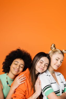 Positive and stylish multiethnic teenage girlfriends with colorful makeup wearing casual outfits while hugging each other with closed eyes isolated on orange, fashionable and trendy clothes clipart
