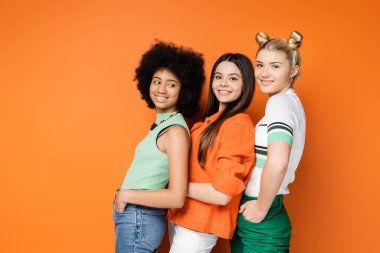 Renkli makyajlı, şık ve çok ırklı genç kızlar el ele tutuşup turuncu arka planda poz veriyorlar, şık ve kendine güvenen pozlar veriyorlar.