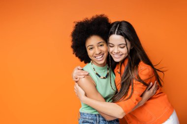 Cheerful and teen african american girl with colorful makeup hugging brunette girlfriend in casual outfit and looking at camera on orange background, stylish and confident poses clipart