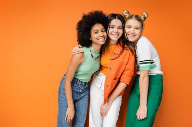 Renkli makyajlı, günlük kıyafetleriyle turuncu arka planda durarak birbirlerine sarılıp poz veren çok ırklı ve çok ırklı genç kızlar şık ve kendine güvenen pozlar veriyorlar.