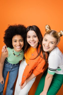 Renkli makyajları ve modaya uygun kıyafetleri olan, turuncu arka planda poz verip kameraya bakarken sarılan çok ırklı gülümseyen genç kızların portresi, moda ve şık saç stilleri.