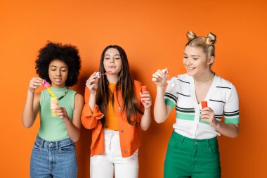 Cheerful blonde teenage girl in casual clothes holding soap bubbles near multiethnic girlfriends with colorful makeup while standing on orange background, trendy and stylish hairstyles clipart