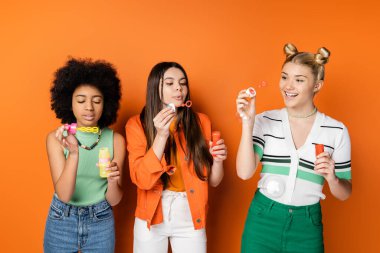 Smiling blonde teenage girl with colorful makeup holding soap bubbles while standing near stylish interracial girlfriends on orange background, trendy and stylish hairstyles clipart